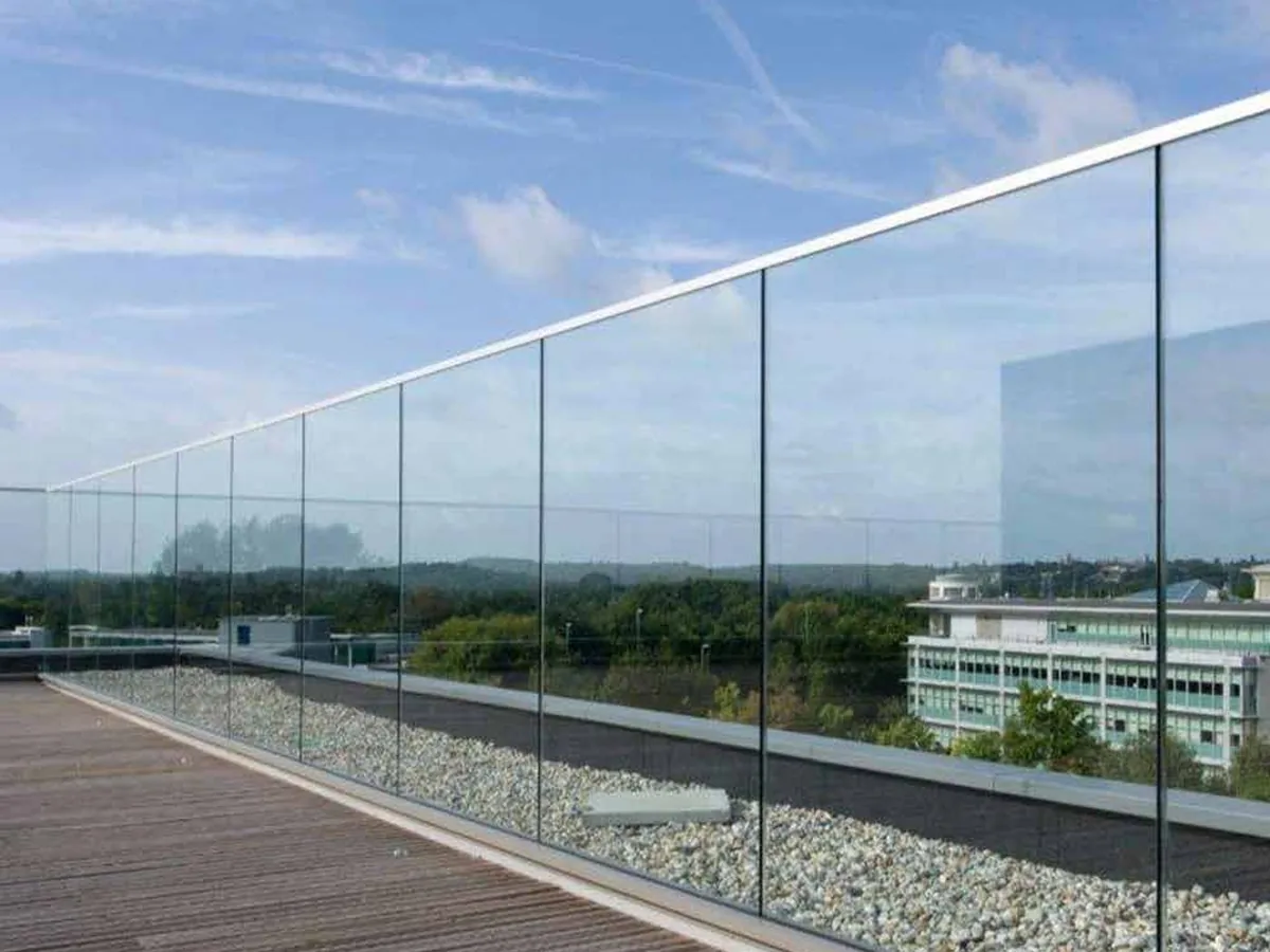 Frameless glass railing with a slim aluminum base shoe running along the edge of a rooftop terrace deck, with open landscape and low buildings beyond.
