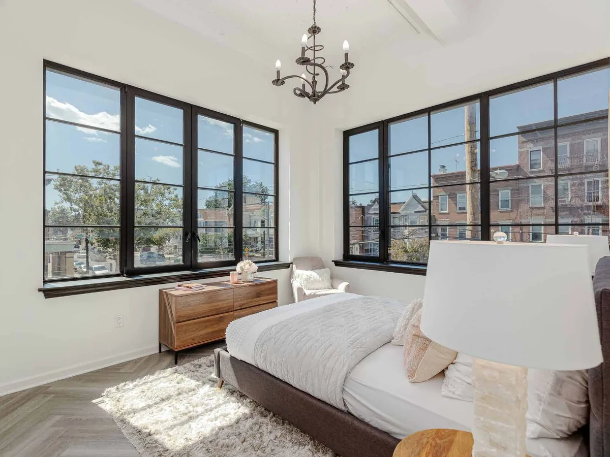 Furnished bedroom with two large dark-framed aluminum windows divided into fixed and operable lights, looking out over rooftops.
