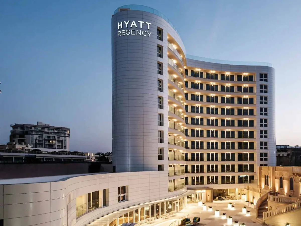 Large hotel elevation at dusk with a continuous glazed and panelled curtain wall grid across several floors, lit from within.