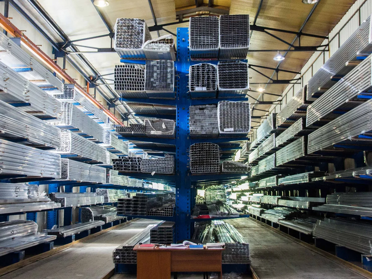 Aisle of tall blue storage racks holding sorted aluminum extrusion profiles inside a production facility.